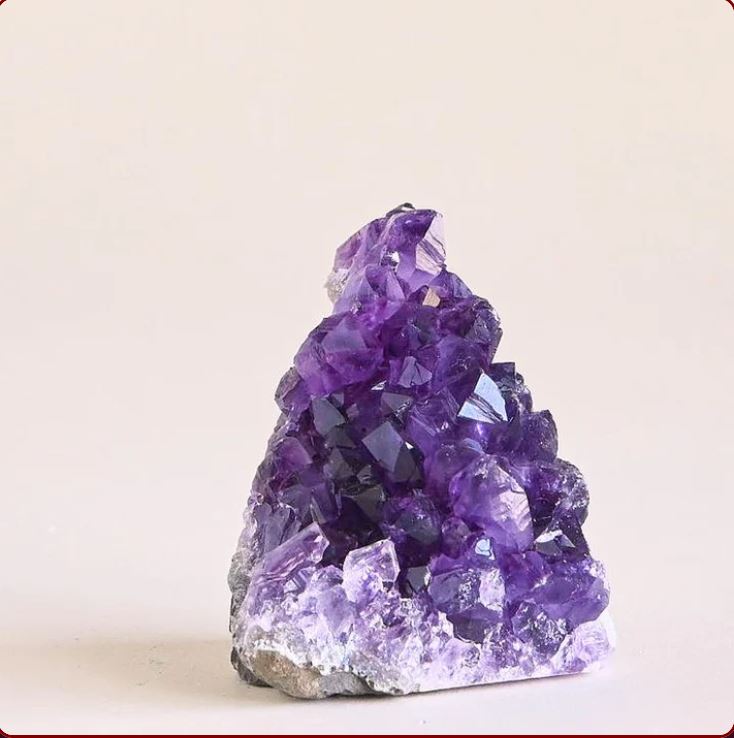 An Amethyst Geode with deep purple crystals, displayed on a white platform against a neutral background. Known for helping with Stress and Anxiety.