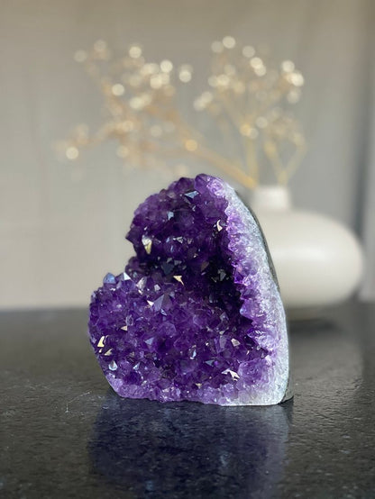 An Amethyst Geode with deep purple crystals, displayed on a white platform against a neutral background. Known for helping with Stress and Anxiety.