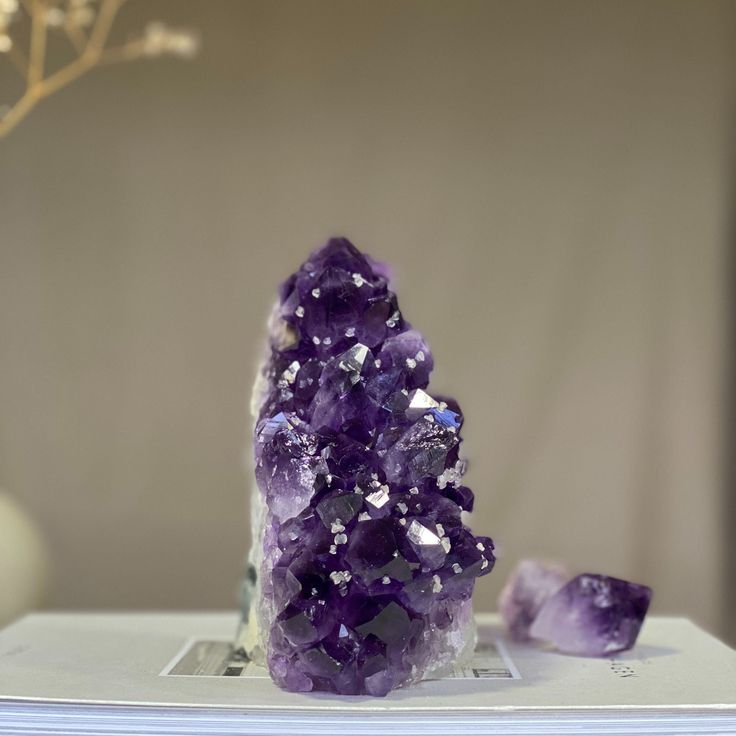 An Amethyst Geode with deep purple crystals, displayed on a white platform against a neutral background. Known for helping with Stress and Anxiety.