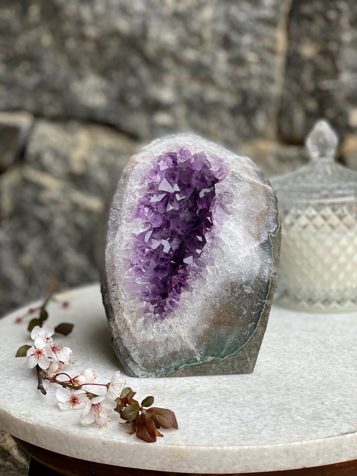 An Amethyst Geode with deep purple crystals, displayed on a white platform against a neutral background. Known for helping with Stress and Anxiety.