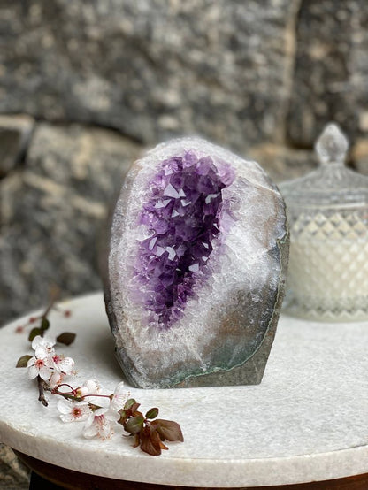 An Amethyst Geode with deep purple crystals, displayed on a white platform against a neutral background. Known for helping with Stress and Anxiety.