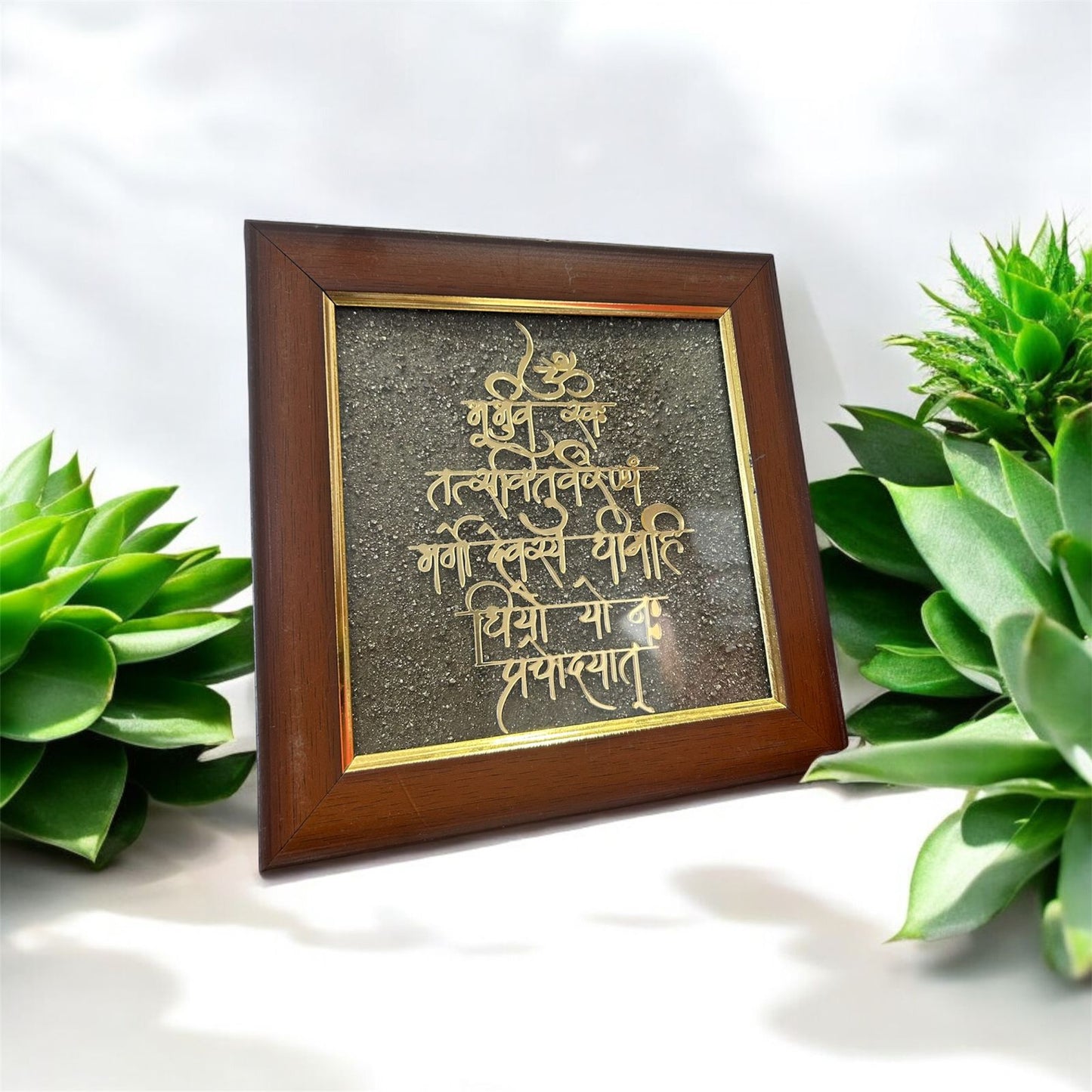 A Pyrite frame with the Gayatri Mantra etched on it, displayed on a white surface with green plants around it. Known for Attracting and peace, this is perfect for office and home decor. 