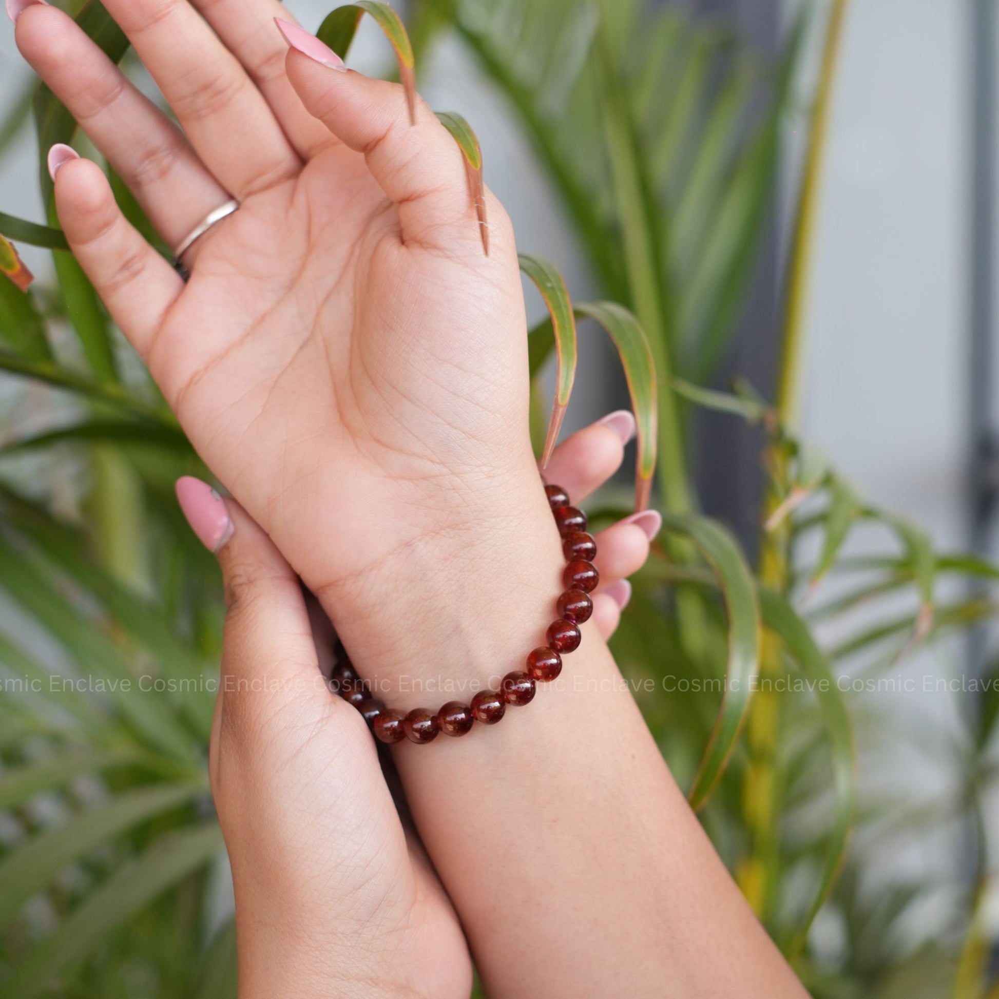 Hand wearing a red beaded Hessonite bracelet with a blurred plant background