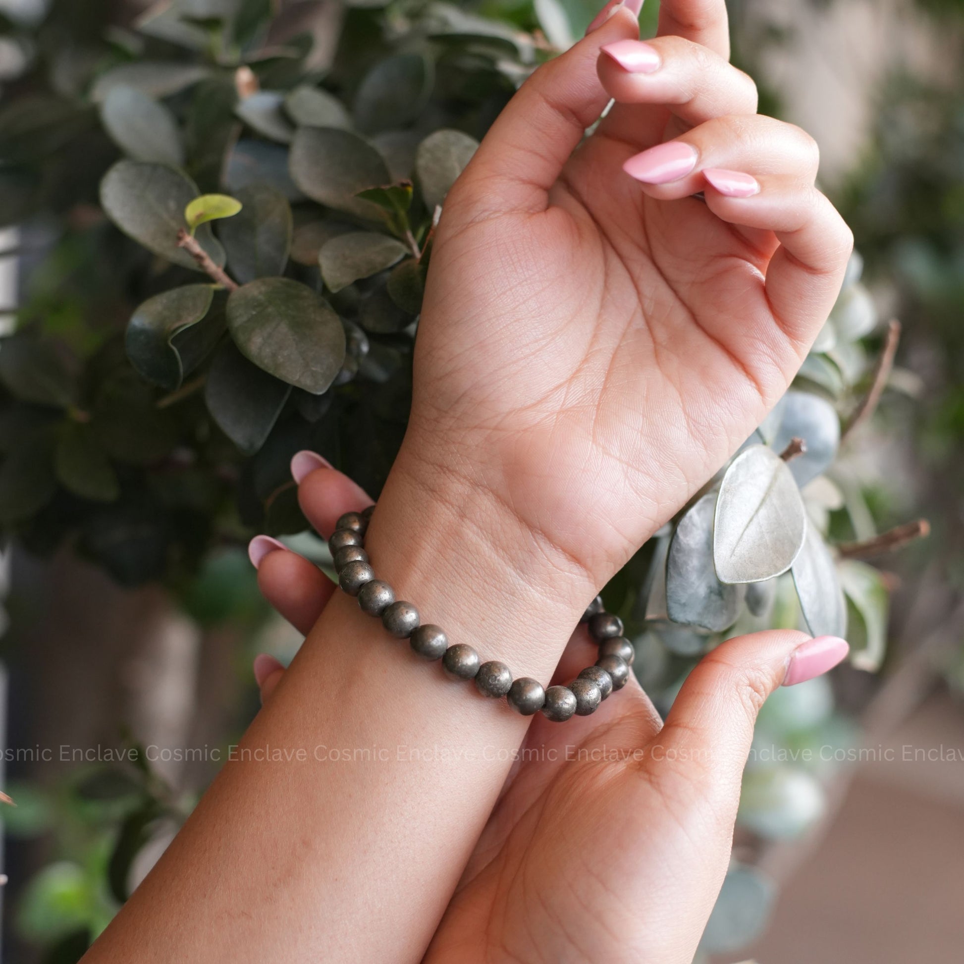 Hand wearing a Pyrite beaded bracelet with a blurred green leafy background
