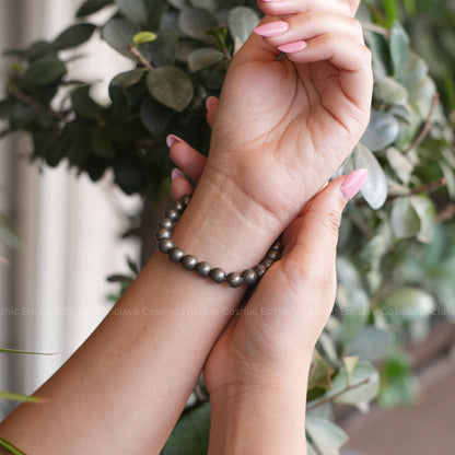 Hand wearing a Pyrite beaded bracelet with a blurred green leafy background