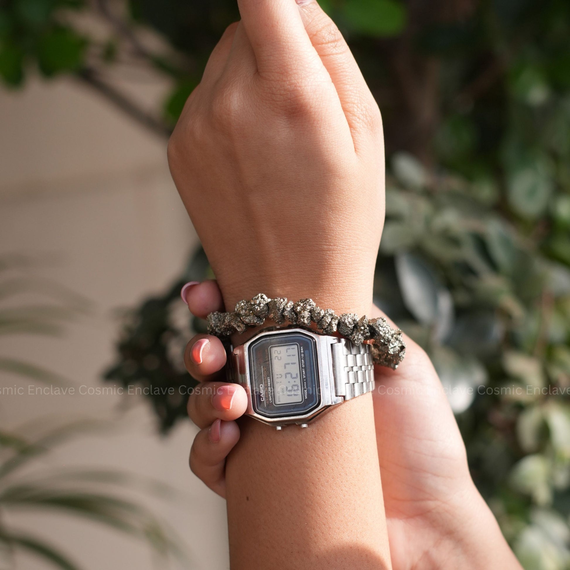 Hand wearing a digital watch with a textured Pyrite Chips bracelet against a blurred green background