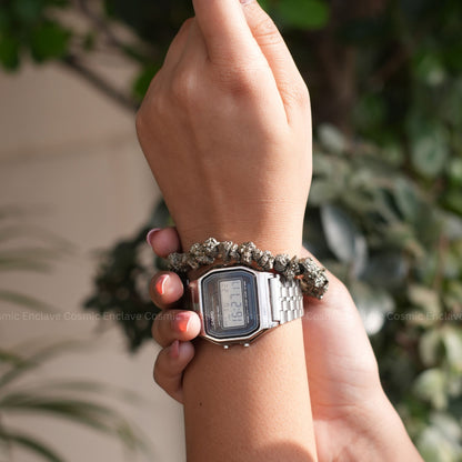 Hand wearing a digital watch with a textured Pyrite Chips bracelet against a blurred green background
