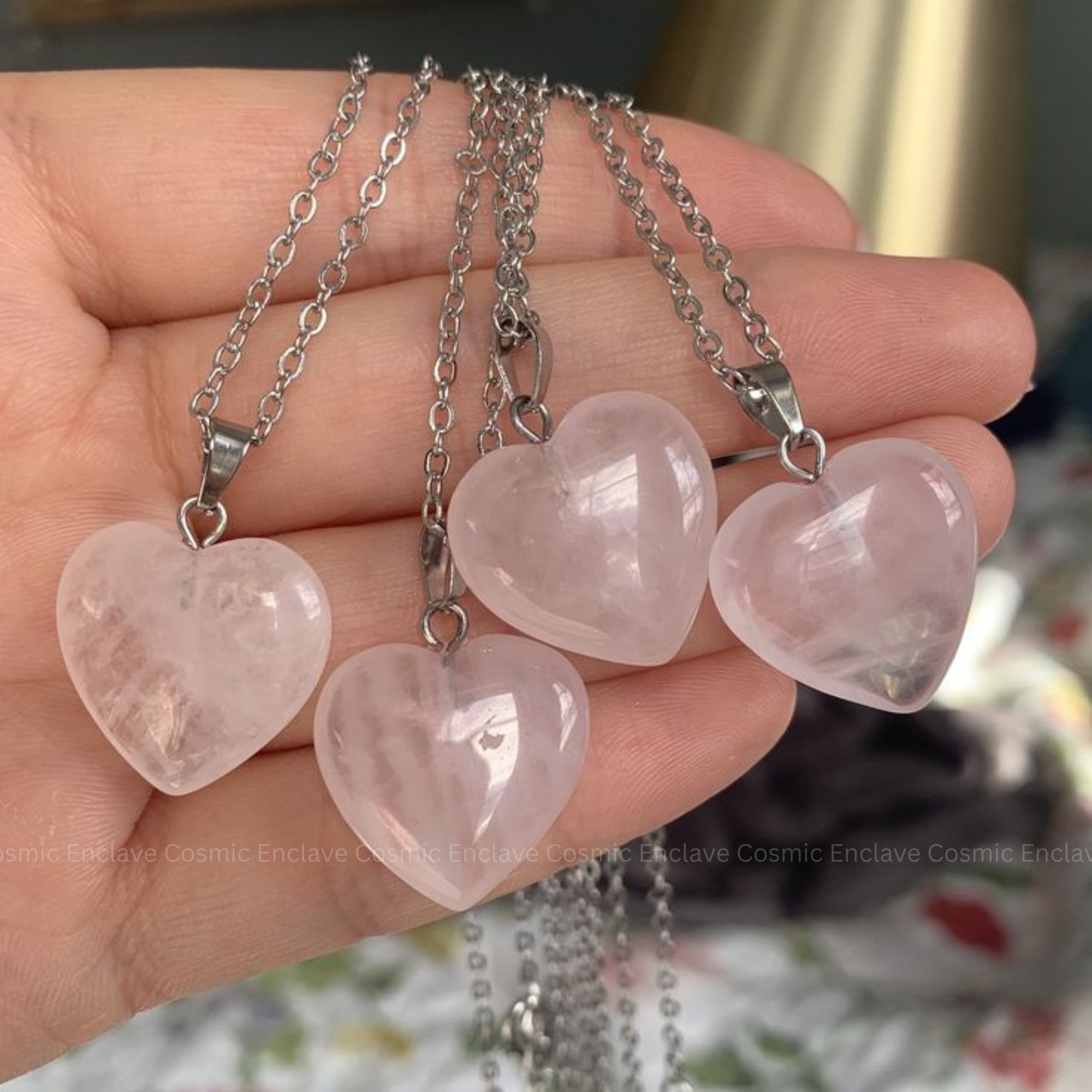 Four heart-shaped Rose Quartz pink stones on silver chains held in a hand with a blurred background.
