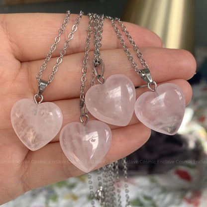 Four heart-shaped Rose Quartz pink stones on silver chains held in a hand with a blurred background.